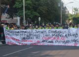 Spanduk sindiran terhadap penyelewengan demokrasi dan represifitas polisi dibentangkan dalam perjalanan menuju titik aksi, Senin (26/08). [BP2M/Anastasia]