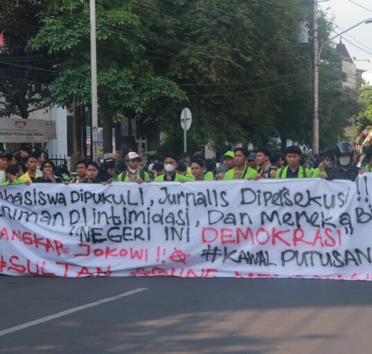 Spanduk sindiran terhadap penyelewengan demokrasi dan represifitas polisi dibentangkan dalam perjalanan menuju titik aksi, Senin (26/08). [BP2M/Anastasia]