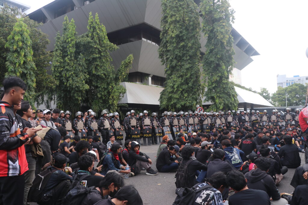 Barisan aparat kepolisian dengan perisai dan helm berjaga di depan massa demonstran yang duduk di halaman Gedung DPRD Jawa Tengah. [Salma/Magang BP2M